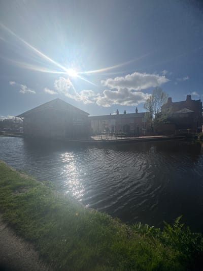 This image depicts a sunny canal scene in Welshpool, United Kingdom. The bright sun, positioned high in the sky to the left, casts a strong lens flare and dazzling reflections across the rippled surface of the canal water.
In the foreground, a vibrant green grassy bank lines the immediate right side of the canal. The canal water itself occupies a significant portion of the mid-ground, reflecting the intense daylight. Across the canal, a row of buildings stands on the far bank. These include several structures that appear to be residential, featuring multiple windows and chimneys.
A prominent, darker brick building further along the canal displays the text "SILVER FISH". To its left, a large, distinctive stone or brick building features the clearly visible text "Y LLANFA". Below this, further text is legible as "Llanfyllin & Llanfair Light Railway Archive, Gallery and Studios," identifying the building's purpose. The sky above is a clear blue with scattered white cumulus clouds.
The scene conveys a bright, pleasant day, likely late morning or afternoon, given the sun's position. No people or specific activities are visible, presenting a serene view of the canal and its surrounding architecture.