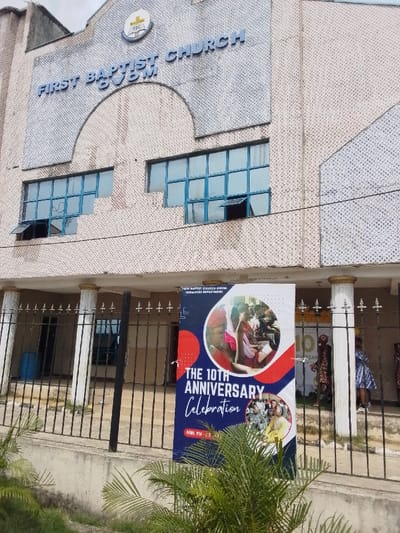 The image shows the exterior of the First Baptist Church, Gwoin, located in Yenagoa, Nigeria, at the coordinates 4.931, 6.268. The church is a two-story building with a white facade, decorated with a patterned design. The front of the building is partially obscured by a black metal gate and a large banner announcing the church's 10th anniversary celebration. The banner features a group of people, likely church members, gathered in an outdoor setting. Several people, including men, women, and children, stand behind the banner, potentially waiting to enter the church. The surrounding area is a green, grassy field with some landscaping. A blue, possibly plastic, barrel rests in front of the church. The building's roof has a slight overhang above the gated entrance.