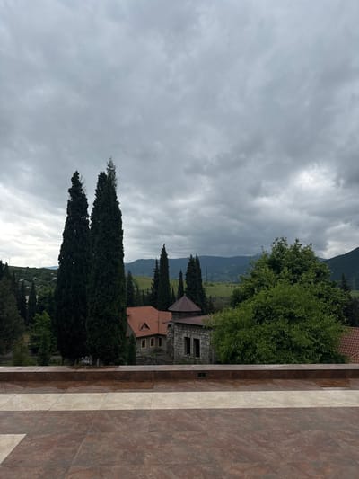 The image depicts a view from a tiled area towards a complex of buildings nestled amongst tall cypress trees and lush green foliage, with rolling hills and a cloudy sky in the background. In the foreground, a wide expanse of large, rectangular tiles in earthy tones of brown and beige forms a walkway or terrace. A low, dark brown tiled wall runs horizontally across the middle ground, separating the tiled foreground from the scene beyond.

Behind the wall, two prominent, slender cypress trees dominate the left side of the frame, their dark green foliage reaching towards the overcast sky. To their right, a stone building with a conical roof and a portion of a red-tiled roof of another building are visible. Further to the right, a large, dense green tree partially obscures a stone building with arched window openings. In the mid-ground, a red-tiled roof with a small triangular dormer window is visible. The background features a landscape of rolling green hills and mountains, with power lines and transmission towers intermittently scattered across the terrain under a heavy, grey, cloudy sky. The overall impression is one of a serene, perhaps rural or semi-rural setting, with a juxtaposition of man-made structures and abundant natural greenery.

It is not possible to definitively confirm the location as Akhtala, Armenia, based solely on the visual information provided in the image.