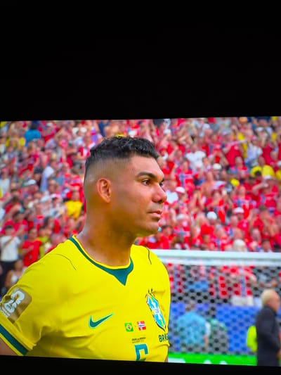 The image shows a close-up of a man in a yellow soccer jersey with green trim. The man is positioned on the left side of the frame, with his head turned to the right, looking out of frame. He has dark, curly hair and a light complexion. The jersey has a blue and green crest on the left breast, a green Nike swoosh on the right breast, and a small Norwegian flag and a Brazilian flag with the text "BRAS" below the crest. A "FIFA" patch is visible on the left sleeve. In the background, a blurred crowd of people wearing red and yellow is visible, suggesting a sporting event. The image is shot from a slightly low angle, looking up at the man. The background is out of focus, with the crowd appearing as a colorful blur. There is no indication of the city of Ognianovo, Bulgaria.