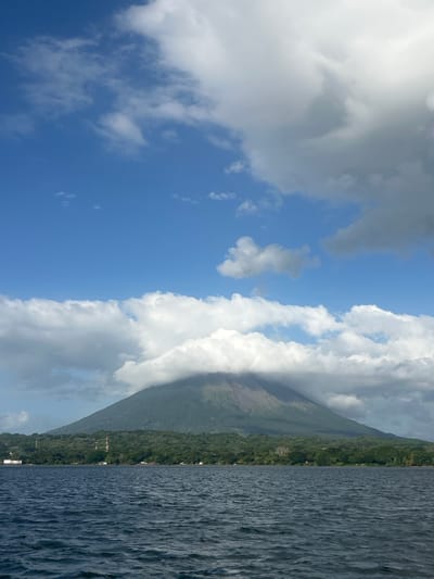 The image depicts a serene natural landscape, with a prominent volcano partially shrouded in clouds dominating the background. The volcano is covered in lush green vegetation, with a visible reddish-brown area near its summit. In front of the volcano, a dense line of green trees forms a natural barrier along the shoreline. The foreground is occupied by a large body of dark blue water, with gentle ripples indicating a calm day. A few structures, possibly buildings and communication towers, are visible on the tree-covered land between the water and the volcano, suggesting a settlement or infrastructure in the area. The sky above is a vibrant blue, interspersed with fluffy white clouds and some darker, more ominous cloud formations. The scene is captured from a low vantage point, likely from a boat on the water, looking towards the shore and the volcano. This scene is characteristic of the landscape around Moyogalpa, Nicaragua, particularly the vicinity of Lake Nicaragua and the Concepcion Volcano.