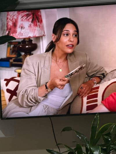 A woman with dark hair pulled back in a ponytail is sitting on a couch, wearing a light-colored blazer and pants. She is holding a small object in her right hand and looking to her left. The couch has decorative pillows with striped patterns. In the background, there is a lamp with a pink floral shade and a bookshelf. The scene appears to be an indoor setting. The location is not discernible from the image.