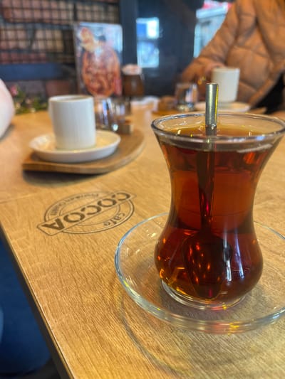 The scene depicts an interior view of a cafe, specifically "COCOA cafe" as indicated by an embossed logo on a light brown wooden table, located in Tekirdağ, Turkey. In the immediate foreground, a clear, tulip-shaped glass is filled with dark amber tea, containing a silver-colored spoon, and rests on a clear glass saucer. To the left and slightly behind, a small white ribbed cup, presumably for Turkish coffee, sits on a white saucer, which in turn is placed on a light-colored, irregularly shaped wooden serving board. The background is blurred, showing a person wearing a light brown padded jacket seated at a table, with another white cup visible behind them. Further in the background, a blurred poster or menu displaying a food item, possibly a pastry or baked good, is visible on a wall, alongside what appears to be a brick-patterned surface. The lighting suggests an indoor setting with both natural and artificial light sources creating reflections on the beverages.