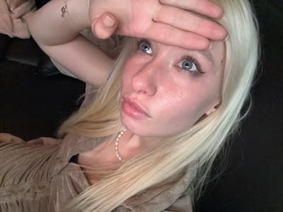 An overhead-angle portrait captures a fair-skinned individual with long blonde hair and blue eyes, located in Namyangju-si, South Korea. The individual is reclined, facing upwards and slightly to the left, with their right hand placed horizontally across their forehead, fingers extended. Their full lips are slightly parted. They wear a light beige or taupe-colored, possibly pleated garment and a pearl necklace. A silver bracelet featuring a key charm is visible on their left wrist. The background is uniformly dark, indicating an indoor setting.