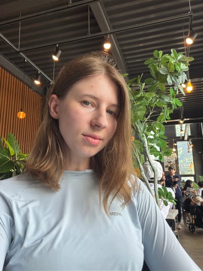 A young Caucasian woman with shoulder-length brown hair and light skin takes a selfie indoors. She is wearing a light blue long-sleeved shirt with the word "NEPA" visible on the chest. The background features an industrial-style ceiling with exposed beams and track lighting, a wooden slat wall, and a large potted plant. In the blurred background, other individuals are seated at tables, suggesting the location is a cafe or restaurant. The scene appears to be in a modern establishment, potentially in Seo-myeon, South Korea.