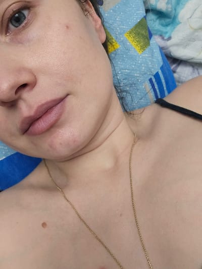 The image shows a close-up view of a person's face and upper chest. The subject has fair skin, blue eyes, and light-colored lips. A gold necklace is visible, draped across the chest. The background consists of a blue and white patterned fabric, likely a pillow or bedding. There are no discernible environmental details to indicate a specific location within Katta Chilanzar-2 MFY, Uzbekistan. The overall scene appears to be a candid, indoor shot.