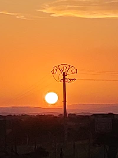 The image captures a sunset in the city of Sobradillo, Spain. A prominent power line pylon stands in the center, silhouetted against the vibrant orange sky. The sun, a bright white orb, is positioned to the left of the pylon, partially obscured by its structure. Several power lines extend from the pylon horizontally across the frame. In the foreground, a dark, silhouetted landscape suggests buildings or natural formations. The sky displays subtle wisps of clouds, illuminated by the setting sun. The overall scene is one of a rural or semi-rural environment during twilight, with the infrastructure of the power line being a dominant visual element against the natural backdrop of the sunset.