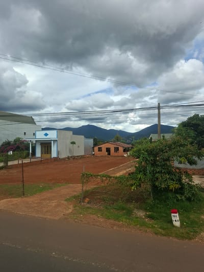 The image depicts a rural or semi-rural landscape under a dramatic, cloudy sky. In the foreground, a paved road runs horizontally. To the right of the road, a white milestone with a red top is visible, marked with "H4 160". Adjacent to the road and the milestone is a grassy area with a chain-link fence.
Further back, a dirt-covered area is prominent, with a fence separating it from the grassy verge. Several structures are visible. On the left, a light blue and white building with a covered entryway and a diamond-shaped emblem above the entrance stands. To its right, a smaller, single-story house constructed of red brick with a tiled roof is situated. In the mid-ground, a large, modern, light-colored building is partially visible on the far left. Dense green foliage and trees are scattered throughout the scene. In the background, a range of dark, mountainous terrain is silhouetted against the sky. The overall impression is a residential area in a region with clear evidence of both development and natural landscape. The scene is identified as being in the city of Tân Phú 2, Vietnam.