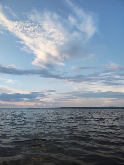 A wide shot captures a calm body of water under a partly cloudy sky. The water's surface is rippled, reflecting the soft light of what appears to be dusk or dawn. In the distance, a thin line of trees marks the horizon, suggesting a shoreline. A small boat is visible on the water, far from the viewer. The sky above is a gradient of pale blue and white, with scattered clouds tinged with hues of pink and orange, indicating either sunrise or sunset. The overall scene is serene and tranquil. The location of Galevo, Russia, is not explicitly depicted or inferable from the visual information provided in the image.