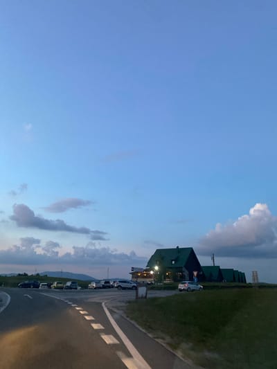 This image depicts a scene at dusk in the vicinity of Javorje, Montenegro. The foreground features a paved road with distinct white lane markings. To the left of the road, a grassy embankment is visible, partially illuminated by an off-camera light source. In the mid-ground, a parking area is populated with several cars, including a dark-colored sedan, a yellow hatchback, and several other vehicles of various colors. Beyond the parking area, a collection of dark green, angular buildings with matching roofs are situated. The largest building, centrally located, appears to be a structure with windows and some exterior lighting. Several smaller, similar structures are positioned in a line behind it. The background consists of rolling hills and mountains under a twilight sky that transitions from a soft blue to darker hues with scattered clouds. The overall atmosphere is serene, indicating the end of the day.
