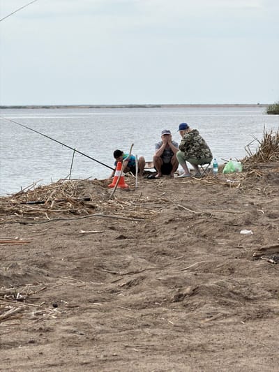 People are fishing on the shore of a body of water near Krasnyi Bor, Russia. The water extends to the horizon under a pale sky. The shoreline is made of sand, dirt, and dead reeds. Two adults are squatting near the water's edge, while another sits on a folding chair a bit further back. A fishing rod is in the water near the adults who are squatting, along with a traffic cone. A bag, a bottle of water, and other small items are on the shore next to the seated adult.