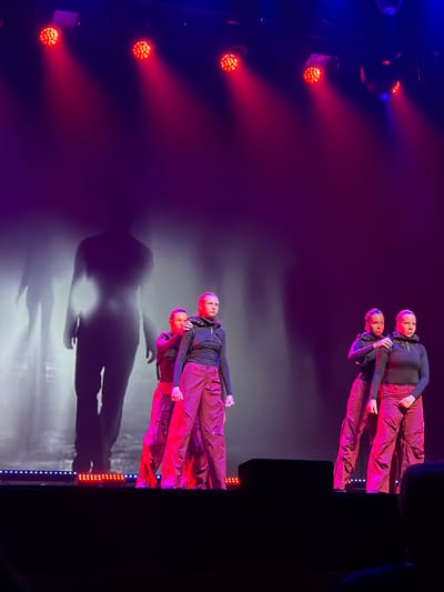 This image captures a stage performance in a venue within Ventspils, Latvia, featuring five female dancers arranged in two groups against a dramatic backdrop. Overhead, multiple red and blue stage lights illuminate the scene, casting conical beams of red light downwards. The background is dominated by large, translucent projections of human silhouettes, appearing faded and ghost-like, with a prominent full-body silhouette centrally located behind the left group of dancers.

On the left side of the stage, three dancers are positioned. The foremost dancer's right side is visible. Behind her, a dancer stands looking straight ahead, hands at her sides, with another dancer behind her placing both hands on the shoulders of the middle dancer, who wears a mesh headpiece.

On the right side of the stage, two dancers are positioned. The front dancer stands slightly angled to the left, looking forward, with her hands down. Behind her, a second dancer has her right hand resting on the left shoulder of the front dancer. Both visible dancers in this group also wear mesh headpieces.

All dancers are uniformly dressed in black long-sleeved tops and loose-fitting, reddish-brown cargo-style pants. The stage floor features a strip of red and blue LED lights along its edge. A dark, rectangular object, possibly a stage monitor or speaker, is visible on the stage floor in the foreground. The overall lighting scheme is dark with strong red and purple atmospheric tones, enhancing the theatrical nature of the performance.