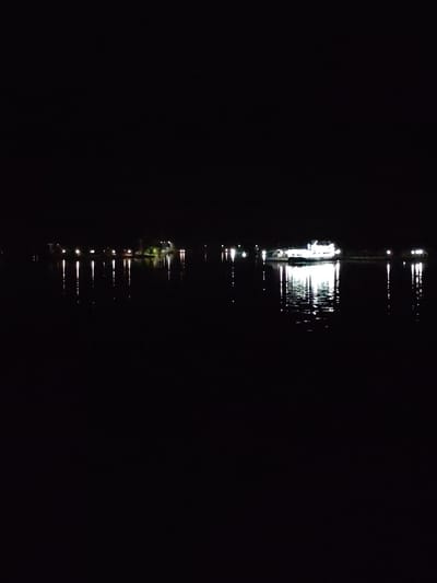 The image shows a night scene in Petrakovo, Russia. A body of water, likely a lake or river, dominates the view. Several lights are reflected on the water's surface. On the left, there are lights that may be from a building or shoreline, along with the reflections. A brightly lit boat is in the center of the image. There are other lights on the far right of the image. The sky is dark and empty. The scene is quiet and still.