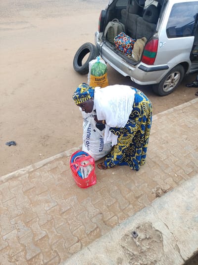 A woman, dressed in a white top and a vibrantly patterned blue and yellow skirt and headscarf, is engaged in an activity near the open back of a silver van. She is bent over, partially obscured by a large white sack. Beside her, also on the ground, is a red plastic bag filled with what appear to be folded items, possibly clothes or fabric. Another large sack, green and yellow, with the word "BANYAN" prominently displayed on it, lies on the ground next to a spare tire. The van's interior is visible, containing several bags and backpacks. The scene takes place on a paved walkway next to a dusty road. The lighting suggests it is daytime. The overall impression is of a commercial transaction or loading/unloading of goods.