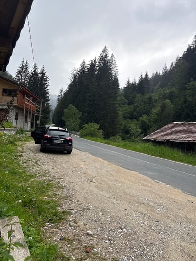This image captures a rural mountainous scene in Здравец, Bulgaria, on an overcast day. A black Infiniti SUV, license plate PB 2204 HE, is parked on a gravel shoulder adjacent to a two-lane asphalt road. The rear driver's side door of the vehicle is open. To the immediate left of the car is a two-story building with a stone foundation and a wooden upper structure featuring reddish-orange accents and a small wooden balcony. Green grass and dandelions grow along the edge of the gravel shoulder on the far left, where a wooden bench or railing is partially visible in the bottom left foreground. A thick black cable or power line extends diagonally downwards from the top left corner of the frame towards the area of the building. The asphalt road, appearing wet, curves slightly to the right, disappearing into the distance. On the opposite side of the road, further right and slightly below road level, an older, smaller building with a tiled roof is partially obscured by tall grass. The entire background is dominated by dense coniferous forests covering steep hills and mountains, receding into a cloudy or misty sky. No individuals are visible in the scene.