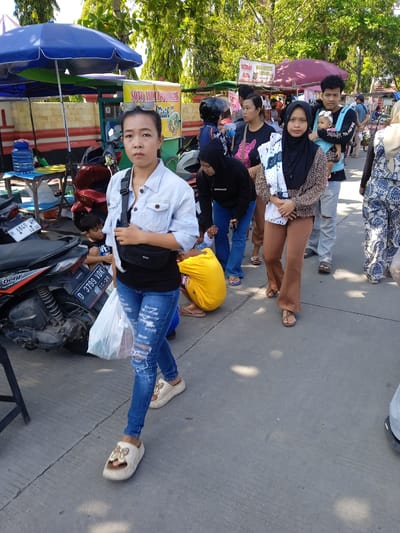 The image captures a street scene in Kebondalem, Indonesia, on a bright, sunny day. A line of people is walking down the street, with a woman in a denim jacket, ripped jeans, and platform sandals in the foreground. She carries a black fanny pack across her chest and a white plastic bag in her hand. Beside her, partially obscured by a parked motorbike, a child is seen sitting behind the bike.

The line of people includes a woman wearing a hijab and holding a blanket, a man carrying a baby in a carrier, and other individuals walking in the same direction. Several food stalls are set up along the side of the road, identifiable by their colorful umbrellas and signage that says "SOTO AMIA BONOVLINE" and "SEKOND". The parked motorbikes indicate the area is accessible by vehicle. The environment appears to be a busy public space, likely a market or shopping area.
