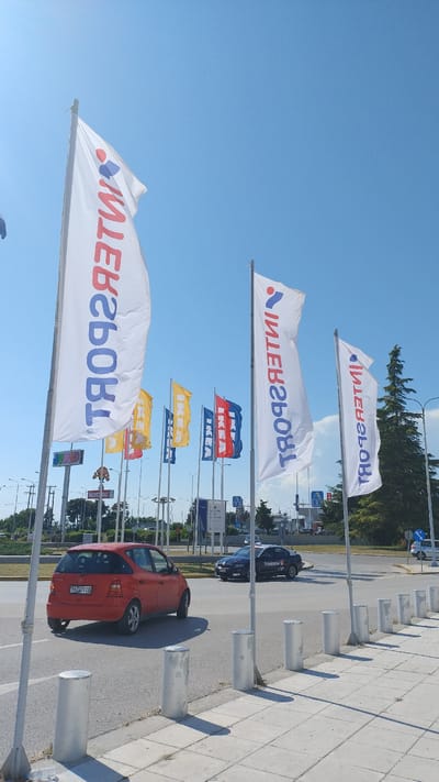 The image depicts a street scene in Pylaia Municipal Unit, Greece. The foreground features a sidewalk with several cylindrical metal bollards. A red Mercedes-Benz A-Class is parked on the left side of the street. In the background, multiple tall flags are displayed, with some featuring the Intersport logo and others displaying the IKEA logo in blue and red. A dark-colored Skoda with the "Freenow" logo is visible driving on the road. The sky is clear and blue, suggesting a sunny day. The environment appears to be an outdoor commercial area, likely near a shopping center or retail park.