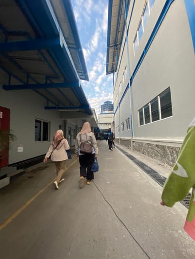 The image depicts an outdoor concrete pathway separating two multi-story buildings within an industrial or commercial complex in Cipeundeuy, Indonesia. The sky overhead is partly cloudy with scattered white clouds.
The building on the left is light grey, featuring a red door, rectangular windows, and a blue-framed awning projecting over the pathway. A yellow line is painted on the concrete alongside this building. The building on the right is also light grey with blue horizontal accents and multiple rectangular windows; its lower section is faced with rough-hewn stone. A dark metal drainage grate runs parallel to this wall along the pathway. In the far background, a taller building with a large water tank on its roof is visible.
Three individuals are walking away from the viewer along the pathway. The leftmost person, a female, wears a patterned light-colored hijab, a light beige jacket, and light brown pants, carrying a dark blue shoulder bag. The central person, also a female, wears a light brown hijab, a grey jacket, dark pants, a dark brown cross-body bag, and carries a dark blue bag in her left hand. A third person, dressed in dark clothing, is visible further down the path. A dark cable or hose lies on the ground, extending from the foreground towards the central figures. A section of an arm covered in green fabric with a white pattern is visible in the extreme right foreground.