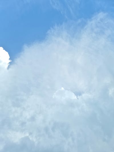 The image is a vertical, full-frame shot of a bright blue sky filled with fluffy white and gray clouds. A large, bright white cloud is prominently featured in the upper left quadrant, with softer, more dispersed clouds filling the rest of the frame. The overall lighting suggests daytime, with the sun likely overhead or slightly to the side, illuminating the clouds. The image does not contain any discernible landmarks or context that would place it specifically within the city of Rzeszów, Poland. There are no people, subjects, objects, actions, or interactions visible in the scene. The environment depicted is solely the sky and clouds.