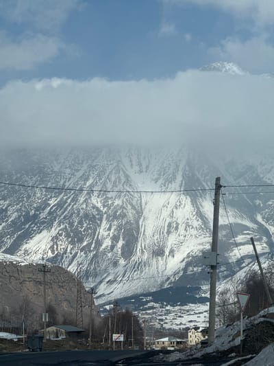 The image presents an outdoor, wide-angle view of a mountainous landscape featuring the town of Sioni, Georgia. The upper portion of the frame shows a blue sky with a prominent, horizontal band of light grey clouds that partially obscure the distant mountain peaks. One snow-capped mountain peak is visible above the cloud line on the upper right.

Dominating the background are vast, rugged mountains extensively covered in snow, with exposed dark rock formations creating a textured surface. In the midground, nestled within a valley, is the settlement of Sioni, Georgia, characterized by numerous buildings with light-colored roofs, interspersed with sparse, bare-branched trees. Patches of snow are visible on the ground throughout the town.

The foreground features a dark asphalt road that curves from the left towards the center-right. Alongside the road, on the left, is a steep, rocky, brownish-grey embankment. Multiple utility poles and lines traverse the scene. A tall, light-colored wooden utility pole stands on the right side of the road, featuring an attached grey electrical box and overhead power lines that extend horizontally across the image. Another wooden utility pole is visible further back on the left, near the rocky slope, alongside a metal lattice power pylon.

On the left side of the road, a small structure with a dark roof and lighter walls is visible next to a dark green/grey wheeled dumpster. A white sign with a red horizontal stripe and a blue sign displaying white Georgian script are positioned further down the road on the left. On the right side of the road, a red and white triangular yield sign is partially obscured by snow and a utility pole. Two residential buildings are visible on the right, one appearing to be under construction with an incomplete roof, and the other a multi-story building with a light-colored facade. Snow patches are present on the ground adjacent to the road and structures.

No individuals, animals, or active vehicles are discernibly present within the image. The scene conveys a cold, winter or early-spring environment.
