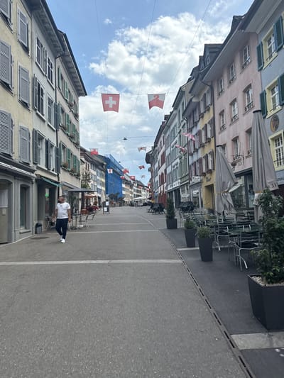 The image shows a street scene in Liestal, Switzerland. The view is looking down a narrow street flanked by buildings on both sides. Buildings have multiple stories and diverse architectural styles, some with window shutters and flower boxes. Swiss flags are strung across the street. A man wearing a white shirt and dark pants is walking along the right side of the street. Tables and chairs are set out along the building fronts, suggesting outdoor dining areas. The sky is visible between the buildings, displaying white clouds against a blue sky. A building is under construction, being wrapped in blue material.