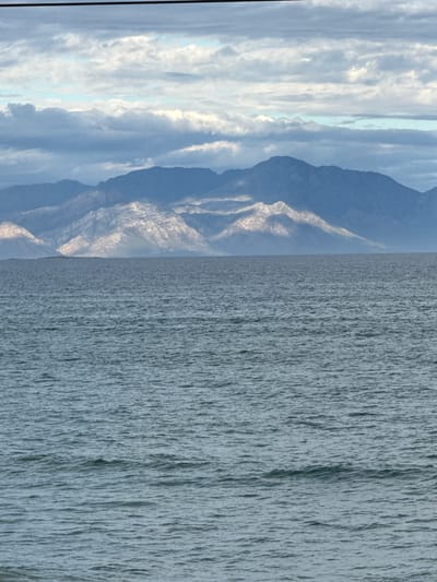 The image shows a landscape featuring mountains on the left side and a large body of water on the right, likely the ocean. The sky is partly cloudy. The mountains are rugged, with visible rock formations and shadows. The water appears choppy, indicated by the vertical lines. The location is possibly Muizenberg, South Africa, given the coastal mountainous landscape, but without explicit confirmation.