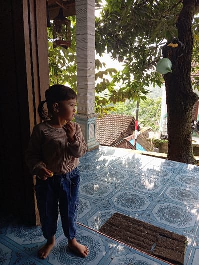 An outdoor scene captured in Bulukandang, Indonesia, shows a young child standing barefoot on a patterned blue tiled porch, partially illuminated by sunlight. The child, wearing a light brown long-sleeved top and blue cargo pants, stands near a wooden door frame on the left, looking rightward. Their left hand is near their mouth, while their right hand holds a small yellow object. To the right, a white fluted column with a light blue base supports the porch roof. Further right, a large tree trunk has a light green plastic watering can and a yellow object affixed to it. In the background, green foliage, the brown tiled roof of a house, and distant hills or mountains are visible under a bright sky. A dark brown rectangular doormat rests on the tiled floor in the lower right. A brown birdcage hangs from the ceiling structure near the wooden frame.