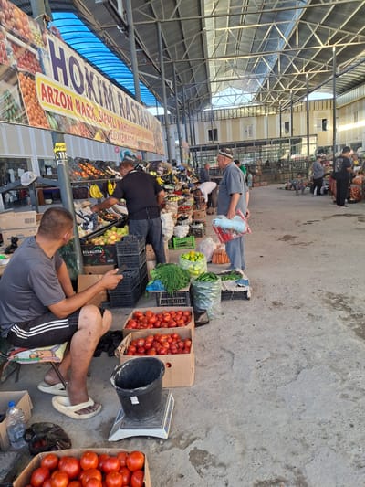 The image shows an outdoor market scene in Ko`kdala, Uzbekistan. A man in a grey t-shirt and black shorts is sitting on a stool, holding a mobile phone and looking down. In front of him are several boxes filled with red tomatoes. To his right, a black bucket sits on a scale. Another man in a black t-shirt and grey pants is standing behind a stall filled with fruits and vegetables, appearing to be serving a customer or arranging produce. The market stalls are set up under a large, open-air roof structure with metal beams and a translucent blue canopy. Various fruits, vegetables, and other goods are displayed for sale. Several other people are visible in the background, some browsing, some working, and some standing and talking. The ground is paved with concrete. A banner with text in a foreign language hangs above the stalls.