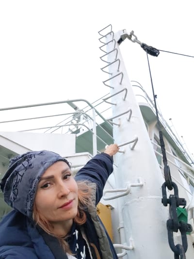 A woman with blonde hair, wearing a blue jacket and a grey patterned headscarf, is pictured from the chest up, looking at the camera. Her right hand is visible on a white ladder attached to a ship's mast. The ship, painted white, has railings and a clear sky above. The image appears to be taken on a boat. The location is identified as Ivanov Bor, Russia.