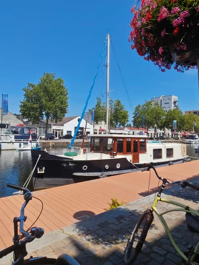 The image depicts a sunny day in Tilburg, Netherlands, likely a harbor area, with a prominent houseboat docked at a wooden pier. In the foreground, two bicycles are parked, one black and one yellow, casting shadows on the cobblestone ground. The houseboat, predominantly black with a white superstructure and wooden accents, has a mast and a blue sail visible. Behind the houseboat, several other boats are moored, and the cityscape includes buildings, trees, and flags. One of the buildings has a sign that appears to read "PINK PALACE MASSAGE." The sky is a clear blue. The scene captures a peaceful urban waterfront environment.