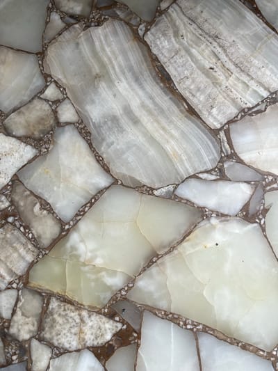 The image shows a close-up of a surface made of irregular pieces of polished, light-colored stone or mineral, possibly marble or onyx, set in a darker-toned matrix of small pebbles or similar material. The stone pieces vary in size and shape, exhibiting swirling patterns and variations in opacity. The matrix filling the gaps between the stones is composed of small, granular particles. There is no indication of the location being Llavallol, Argentina, nor any people, objects, actions, or interactions.