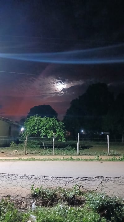 The image depicts a night scene in El Samán, Venezuela. A road runs horizontally across the frame. To the left of the road is a field with a cloudy night sky and a bright moon. Trees are visible in the background, silhouetted against the sky. To the right of the road, there is a wire fence with vegetation growing on either side. Wooden posts are visible along the road and fence line. There are no people visible in the image. The scene is illuminated by the moon and possibly streetlights.