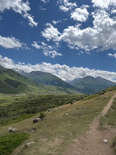This is a landscape photograph taken from a dirt path on a grassy hill. The path curves from the bottom right corner of the frame upwards and to the left. The hill is covered in dry grass and scattered large grey rocks. In the middle ground, there is a valley with green, rolling hills, a road, and a small village. The background features several large, green mountains, with some snow-capped peaks visible on the tallest ones. The sky above is a bright blue with scattered white cumulus clouds. The image appears to be taken in the vicinity of El'tiubiu, Russia, a mountainous region. There are no people or animals visible in the frame.