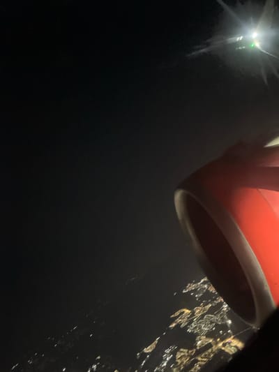 From a high vantage point, a city sprawls into the night, its lights scattered like gems on a dark cloth. A red and white object, possibly a mug or container, dominates the upper portion of the frame, its rounded edge creating a visual foreground. A distant tower emits a strong light, contrasting with the pervasive darkness. The scene evokes a sense of distance and perspective, contrasting the human element (the object) with the vastness of the urban landscape.