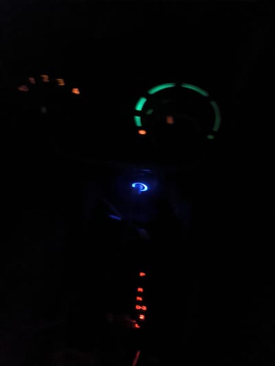 The image shows the interior of a vehicle at night. Illuminated gauges are visible: a tachometer with green markings and an indicator with orange numerals 1-4. A blue-lit button is located lower in the center of the image. Beneath the button, a gear selector indicator displays the letters P, R, N, D, 2, and L in red. The gear selector appears to be in the "L" position. The location in Keudealue Ie Puteh, Indonesia is not visible from the image.