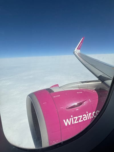 This image presents an aerial view from an airplane window, with the location context given as Ashnak, Armenia. The main subjects are the aircraft's bright pink engine and a portion of its wing, framed by the dark gray outline of the window on the right.

The engine casing is predominantly pink, boldly displaying "wizzair.com" in large white letters across its side. Several warning labels, reading "NO STEP" in white text on a reddish background, are visible on the engine, some accompanied by directional arrows. The large air intake of the engine is also visible towards the bottom right of the frame.

Extending from the left side of the frame, the airplane wing is partially visible, featuring a distinct pink winglet at its tip, which also carries the "wizzair.com" branding in white text. Below the plane, a thick layer of white clouds or haze stretches across the horizon, indicating the aircraft is flying above a significant weather layer. Above the clouds and to the left of the wing, the sky is a clear and deep blue. The bright sunlight reflecting off the engine and wing suggests it is daytime. The image captures the serene yet powerful essence of high-altitude flight. No individuals are visible in the photograph.