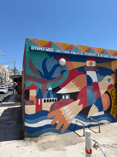 In Gżira, Malta, a vibrant mural adorns the side of a building, situated on a street with moderate traffic. The mural, painted in bold, abstract shapes and colors, features a lighthouse, birds, waves, and architectural elements like domes. Along the top edge of the mural, white lettering reads "REFUGEE WEEK. UNITING. MALTESE. MIGRANT. & REFUGEE. COMMUNITY."
The building with the mural is positioned at a corner, with a sidewalk running alongside it. The sidewalk is paved with stone slabs and has a metal drain cover. A white street lamp is visible in the background to the left. Further down the street, there are several multi-story buildings, a bus stop shelter, and vehicles, including a white car.
In the foreground, a white post with red markings stands near the mural, secured by a chain and a padlock. Two metal racks, likely for bicycles, are attached to the building near the base of the mural. The sky is clear and blue, indicating a sunny day. The overall scene depicts a public art installation in an urban environment.