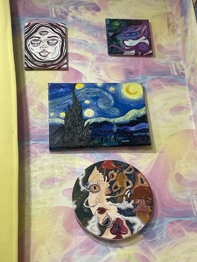 This is a close-up, eye-level, vertical shot of four small, framed paintings displayed on a wall covered in a psychedelic, swirled pattern of pastel pink, yellow, and purple. A bright yellow curtain or fabric hangs vertically on the left side of the frame, obscuring a portion of the wall.
The paintings are arranged in a staggered, somewhat haphazard fashion.
At the top left is a small rectangular painting featuring a black and white line drawing of a face with three eyes and swirling lines around the head in dark purple.
Above and to the right of the central painting is another small, square, abstract painting with purple, blue, and green colors, featuring what appears to be a stylized creature or organic shapes.
Dominating the center of the image is a rectangular painting of Vincent van Gogh's "Starry Night." It depicts a dark, jagged cypress tree in the foreground, a village with buildings and a spire in the middle ground, and a swirling night sky with a crescent moon and bright yellow stars.
Below the "Starry Night" painting is a circular painting with a surreal, psychedelic design. It features a face with large, wide eyes, red lips, and is surrounded by mushrooms and abstract shapes. The background of this painting is dark blue with scattered colorful elements.
The overall impression is of an eclectic art display in a brightly decorated, possibly bohemian or artistic, living space. The specific location within ЖК «Борисоглебское», Russia, cannot be determined from the visual information provided in the image.