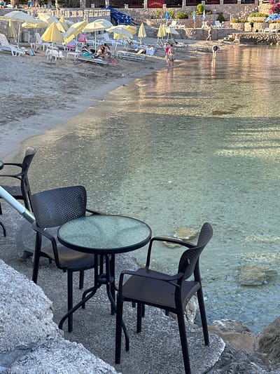 The image displays a coastal scene in Seoca, Montenegro, under warm, low-angle sunlight. In the immediate foreground, a round table with a textured glass top is positioned between two dark-colored plastic chairs, each with a perforated backrest. This furniture rests on a grey, pebbled concrete surface situated at the very edge of calm, clear, shallow water. An irregular grey rock structure borders the concrete on the left.
The water, extending from the foreground, reveals a light-colored, rocky bottom and reflects sunlight, creating shimmering patterns. To the left of the water, a light-tan sandy beach is occupied by numerous open yellow and white beach umbrellas, beneath which multiple white plastic lounge chairs are arranged.
Several individuals are present within the scene. In the shallow water, one person wearing a dark swimsuit is bent over, facing away from the viewer. On the sandy edge, two individuals stand facing each other: one in a pink swimsuit and another in a patterned dress. Approximately 5-8 additional people are indistinctly visible relaxing on lounge chairs under the beach umbrellas.
In the background, a multi-tiered stone structure with green foliage ascends from the beach. A building on the uppermost tier features a red sign prominently displaying the word "Cocktails." Additional people are discernible walking or standing on these higher terraces.