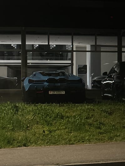The image depicts a blue BMW M4 parked next to a modern building with glass windows in Kloten, Switzerland. The license plate reads "ZH-4004". The car is positioned alongside a patch of green grass, with a sidewalk adjacent to the grass. Another dark-colored vehicle, potentially a BMW, is parked in front of the blue BMW. The overall setting appears to be a car dealership.