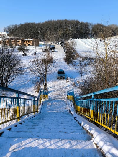 The image displays a snow-covered winter landscape in Prybuzke, Ukraine, under a clear blue sky. In the foreground, a set of outdoor steps, coated with snow, descends into the scene. The metal handrails on either side are painted in alternating blue and yellow stripes, indicative of the Ukrainian flag colors. Below the stairs, a light-colored van is parked on a snow-covered path, facing away from the viewer. To the left, several multi-story brick buildings, appearing to be under construction, are visible. An all-terrain vehicle (ATV) and a few individuals are present on the snow in front of these buildings. Further into the background, a recreational snow tubing or skiing slope with multiple parallel tracks is visible on the right, populated by several individuals. Numerous parked vehicles line a snowy road traversing the scene towards the base of the slope. A dense forest of bare, deciduous trees forms the distant background.