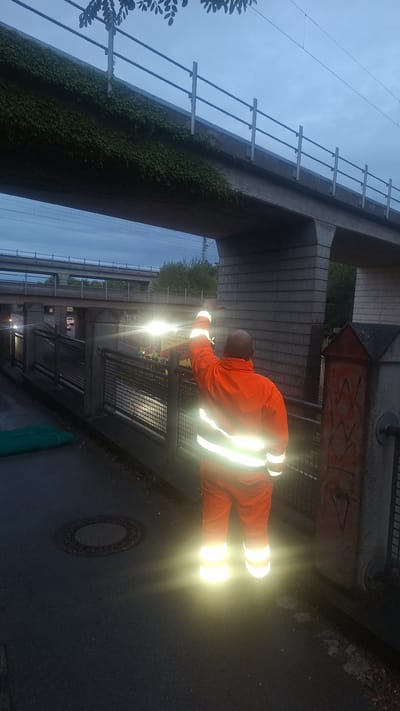A person in a bright orange high-visibility suit with reflective stripes is standing and pointing with their right arm raised. The person is facing away from the camera, towards a busy street or highway overpass. There are bright lights from vehicles in the background, suggesting it is either dusk or nighttime. The person is standing on a paved area next to a metal fence, below a large concrete overpass that has ivy growing on its side. In the foreground, there is a circular metal manhole cover on the ground. The overall scene appears to be an urban environment, likely an industrial or transportation infrastructure area, given the overpass and road.