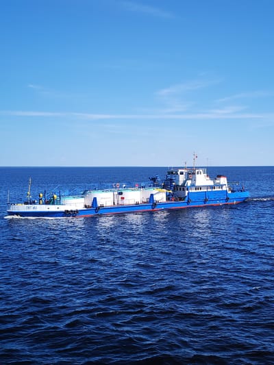 A blue and white specialized vessel, identified by the Cyrillic inscription "ПНТ 451" on its bow and "ОГНЕОПАСНО" (FLAMMABLE) on a white tank, transits a large body of deep blue water. The vessel features a blue hull, a white superstructure, and a red waterline stripe. Cylindrical white tanks are visible on its forward deck. It is actively moving from left to right, generating a visible wake. The water surface exhibits ripples. The sky is clear light blue with a few faint wispy white clouds. The horizon line is distinct, with no landmass visible in the distance. The scene is illuminated by bright daylight. This event occurred in Мякса, Russia.