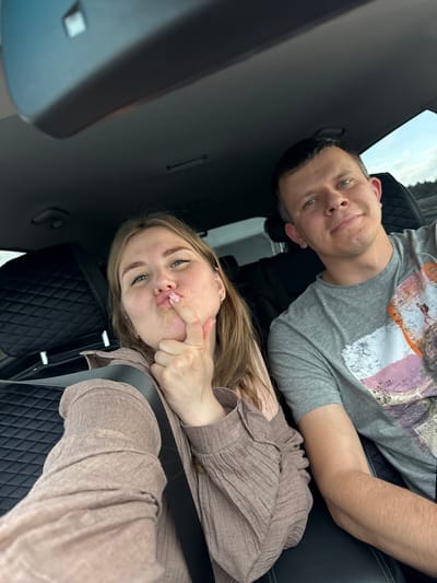 This media file is a portrait of two people, a man and a woman, who appear to be sitting in the back seat of a car. The man is on the left, wearing a grey t-shirt with a colorful graphic print. He has short dark hair and blue eyes and is looking towards the camera with a slight smile. The woman is on the right, with long blonde hair, and is wearing a light brown, long-sleeved shirt. She is making a pouting face with her finger on her lips, and she also has blue eyes. Both individuals appear to be in good spirits, with the woman's playful expression suggesting a lighthearted mood. The interior of the car is visible, with dark, quilted leather seats and a seatbelt. The overall lighting suggests it might be daytime, possibly overcast. The location is identified as Kasimovo, Russia.