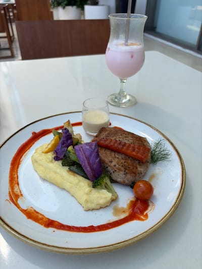 A plate of food is presented on a white surface, presumably a table. The meal consists of what appears to be a seared piece of meat topped with an orange slice, garnished with dill. Alongside, there's a tomato, a sauce, puréed potatoes, and a medley of vegetables, including purple cabbage, zucchini, and broccoli. A glass of a light pink beverage and a small glass of a white sauce sit next to the plate. The scene appears to be indoors, possibly a restaurant in Talpe, Sri Lanka, based on the prompt. The image is taken from a slightly elevated angle, showcasing the arrangement of the food on the plate. The setting is simple, with minimal visible background details, focusing attention on the food presentation.