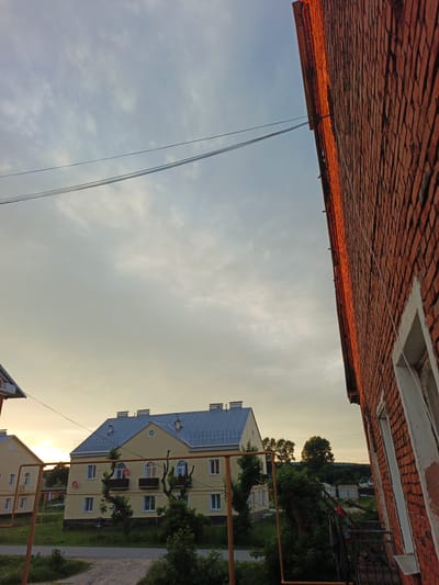 The image depicts an outdoor scene in Medvedyovka, Russia, at dusk. The sky is overcast with a pale yellow hue near the horizon, indicating the sun is setting. In the foreground on the right, a wall of red brick is prominently featured, with a white-trimmed window visible. The brickwork shows texture and detail. On the left, two yellow apartment buildings with gray, sloped roofs are visible. These buildings have multiple windows and balconies. Orange metal poles, likely part of a fence or support structure, are present in front of the buildings. Trees and vegetation surround the buildings, suggesting a residential area. Power lines are strung across the sky, adding a man-made element to the natural landscape. The overall mood is calm and serene, typical of an evening in a rural or suburban setting. There is no visible text in the image.
