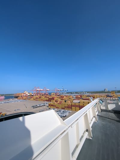 This is a high-angle, wide shot of a bustling port under a clear blue sky. The foreground is dominated by the deck and white railing of a ship. In the mid-ground, a vast expanse of stacked shipping containers, primarily in yellow and red, fills the scene. Several large, pink gantry cranes are positioned amongst the containers, indicating active cargo operations. Beyond the containers, a large cargo ship is docked, also laden with containers. Further in the background, a large parking lot filled with many white cars is visible, suggesting a car terminal within the port. The distant background shows a coastline with sparse vegetation and a tall, light-colored building. The port appears to be the Hambantota International Port in Hambantota, Sri Lanka, a location known for its extensive container and vehicle handling facilities. There are no people clearly visible in the image.