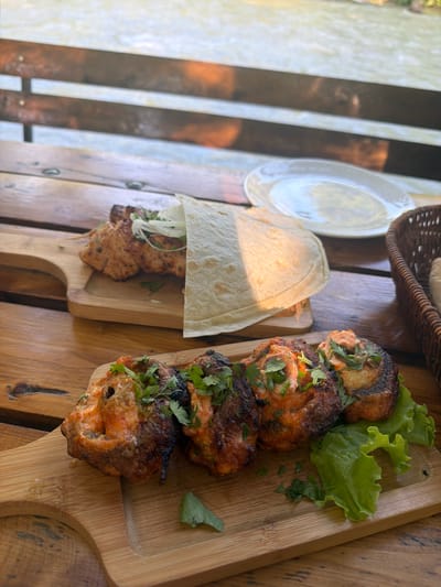 Two wooden platters of food are on a wooden table outdoors, with a river in the background. The platter in the foreground contains four pieces of grilled meat topped with a creamy sauce and chopped cilantro, placed on a bed of lettuce. The platter in the background holds grilled chicken skewers wrapped in flatbread and topped with onions. A white plate and a woven basket are also visible on the table. The setting suggests a restaurant or dining area near a natural body of water, potentially in Abzhanda, Abkhazia.