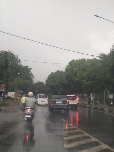 This is a vertical, eye-level shot of a rainy street scene in Meunasahmanyang, Indonesia. The weather is overcast, and the road is wet and reflective, with visible water droplets on the lens. A person on a scooter, wearing a light green jacket and a pink helmet, is in the foreground, facing away from the camera and traveling on the wet road. The scooter has a license plate that reads "BL 5775 LAJ".

Several cars are also on the road. Behind the scooter is a white car, and further ahead, a black SUV and another white car. The reflections of brake lights are visible on the wet surface of the road. The scene is lined with lush green trees on both sides of the road, with a few power lines running overhead. There are also streetlights visible in the background.

On the left side of the road, there are buildings and a white van. Some text is visible on a red sign to the left, partially obscured but appearing to say "NEW". The overall mood is damp and hazy due to the rain. The time of day appears to be late afternoon or early evening, given the dim, overcast sky.