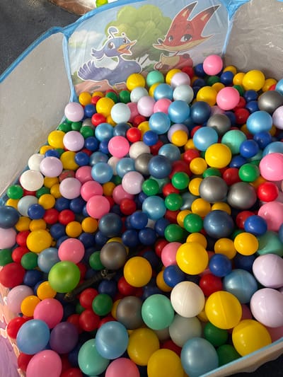 A high-angle, close-up shot displays a ball pit filled with numerous colorful plastic balls. The balls are predominantly shades of blue, yellow, pink, red, green, and white, with some silver and gray ones mixed in. The ball pit appears to be a soft-sided playpen, with a printed backdrop featuring cartoon characters of a blue bird and a red fox against a green, leafy background. The ball pit is located indoors, on a dark, textured surface, possibly carpet or rubber matting, in the city of Благовское Снт, Russia. There are no people visible in the image.